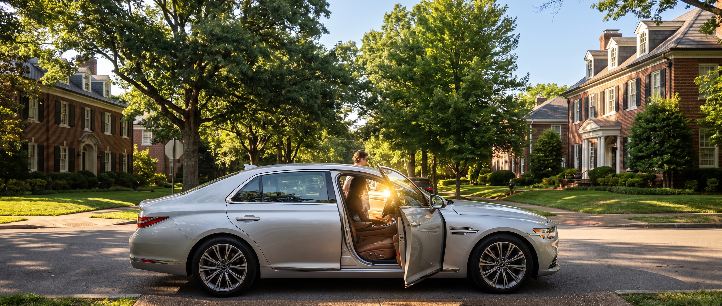 2026 Genesis G90 sedan in silver, parked on a tree-lined residential street in an affluent Nashville neighborhood on a warm summer afternoon, driver's door open with sunlight streaming through
