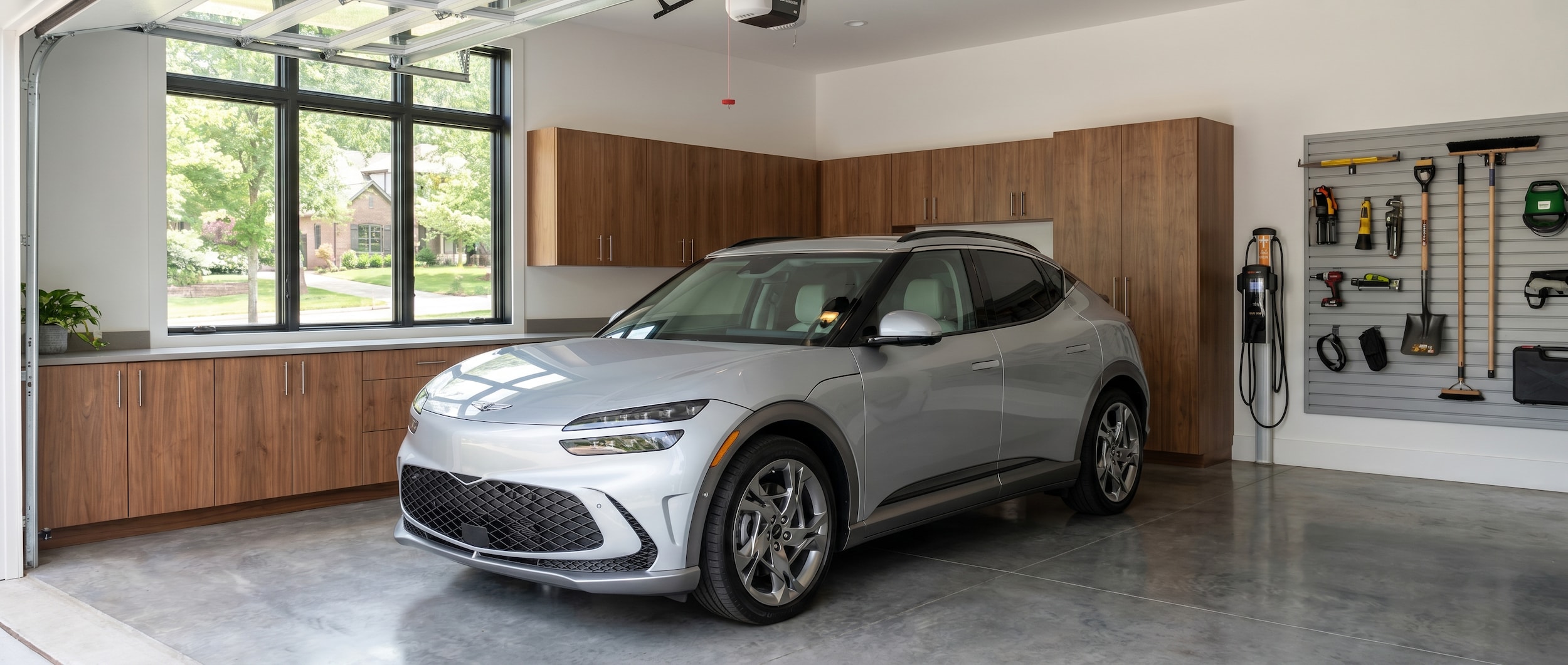 A silver Genesis GV60 parked in a clean, modern suburban garage in Brentwood, Tennessee, with a Level 2 EV charger mounted on the wall, warm interior lighting suggesting an upscale home setting.
