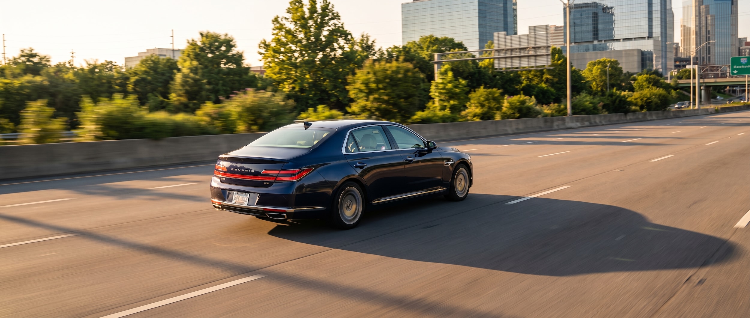 A 2026 Genesis G90 in a refined dark exterior color moving along a sunlit Nashville urban highway corridor, late afternoon light casting long shadows across the road, green summer foliage visible along the roadside, dashboard softly lit inside the cabin