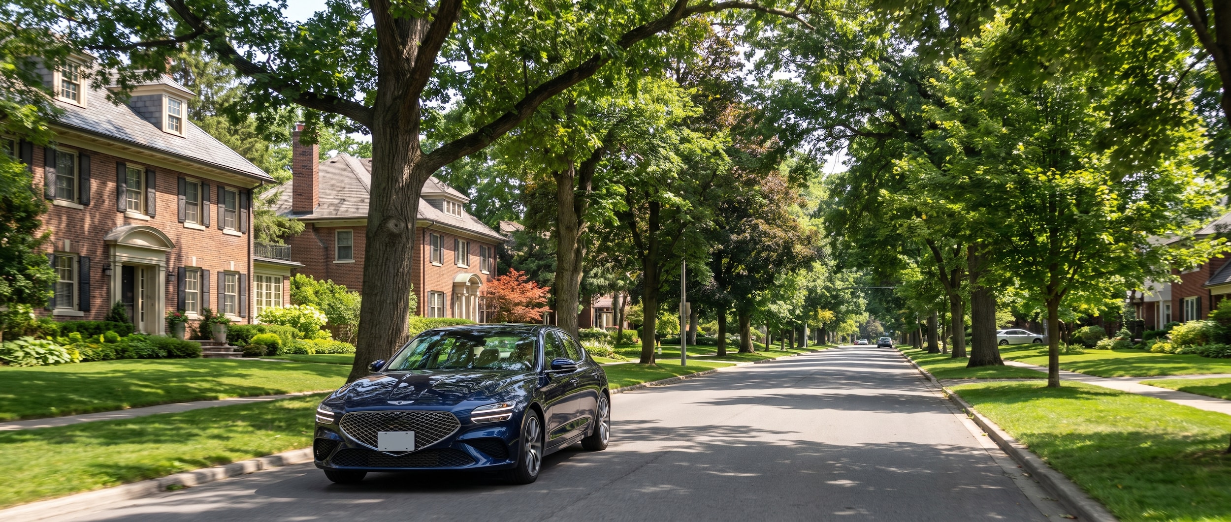 A 2026 Genesis G70 in a deep blue finish driving along a wide, tree-canopied boulevard lined with mature hardwoods and manicured estates, evoking the residential streets of Belle Meade, Nashville, in summer daylight.