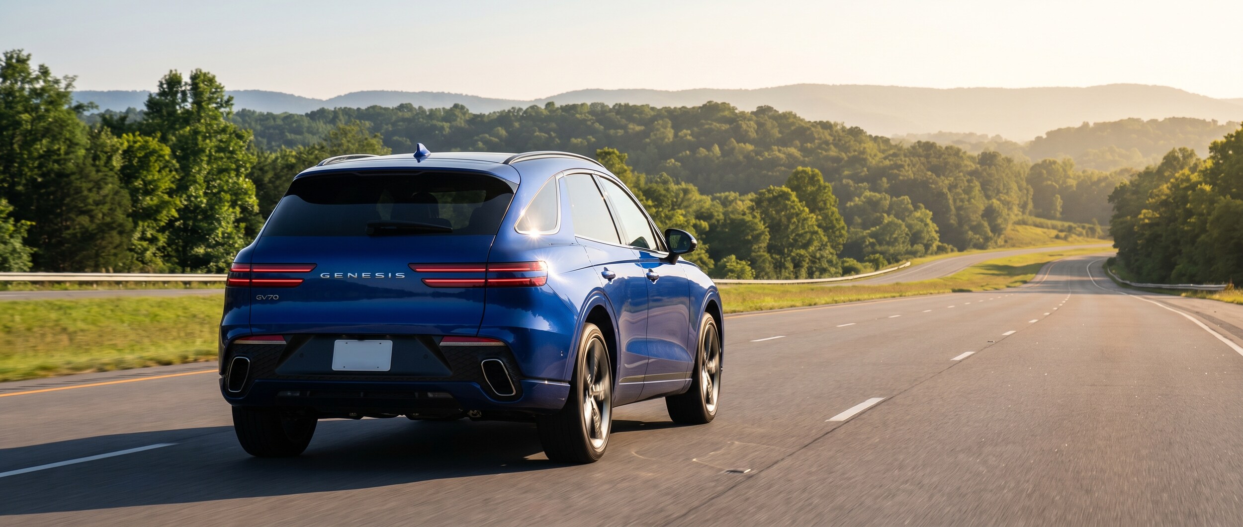 A 2026 Genesis GV70 in a warm silver or Matira Blue finish cruising a sunlit Tennessee highway with tree-covered rolling hills in the background, summer haze visible on the horizon, shot from a low three-quarter rear angle that shows the distinctive two-line taillights
