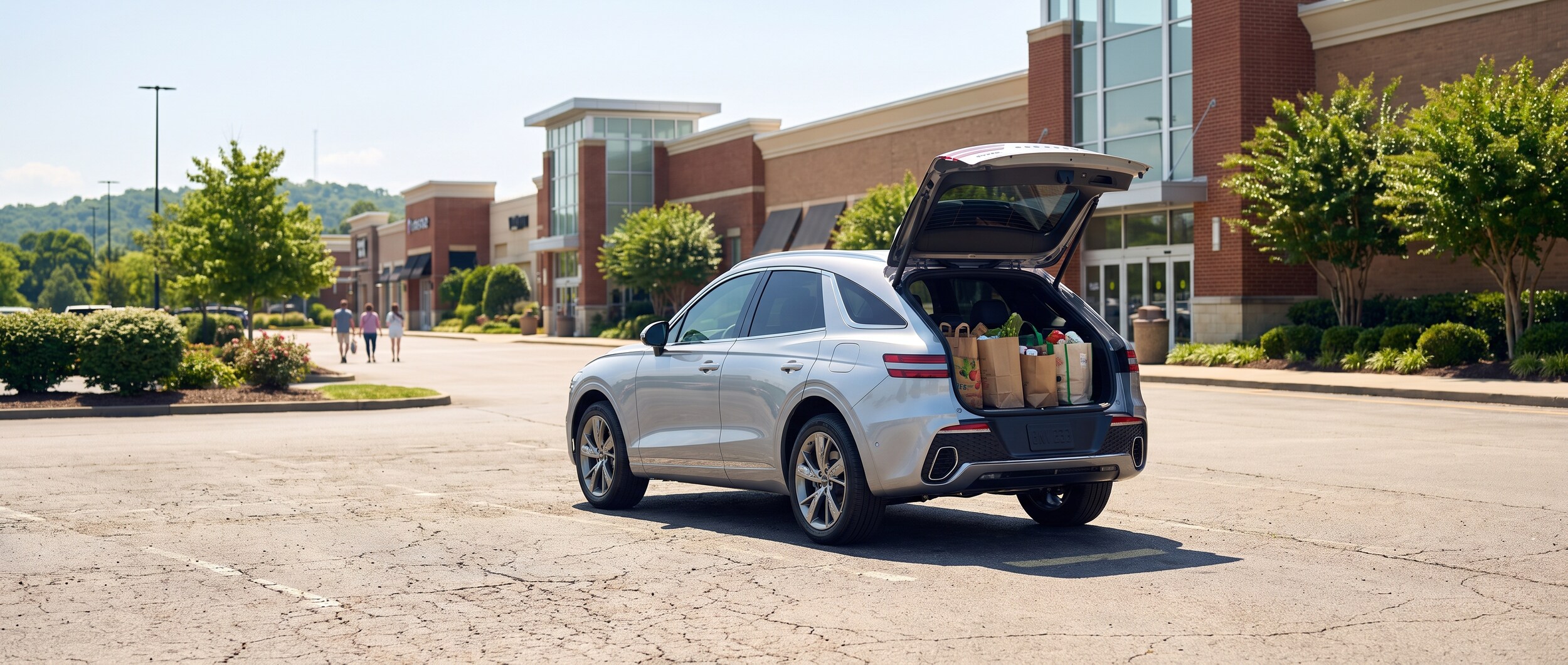 A 2026 Genesis GV70 in a bright silver or white finish parked in a sun-lit open surface lot in front of a large suburban shopping mall in summer, rear hatch open with shopping bags inside, Williamson County Tennessee context