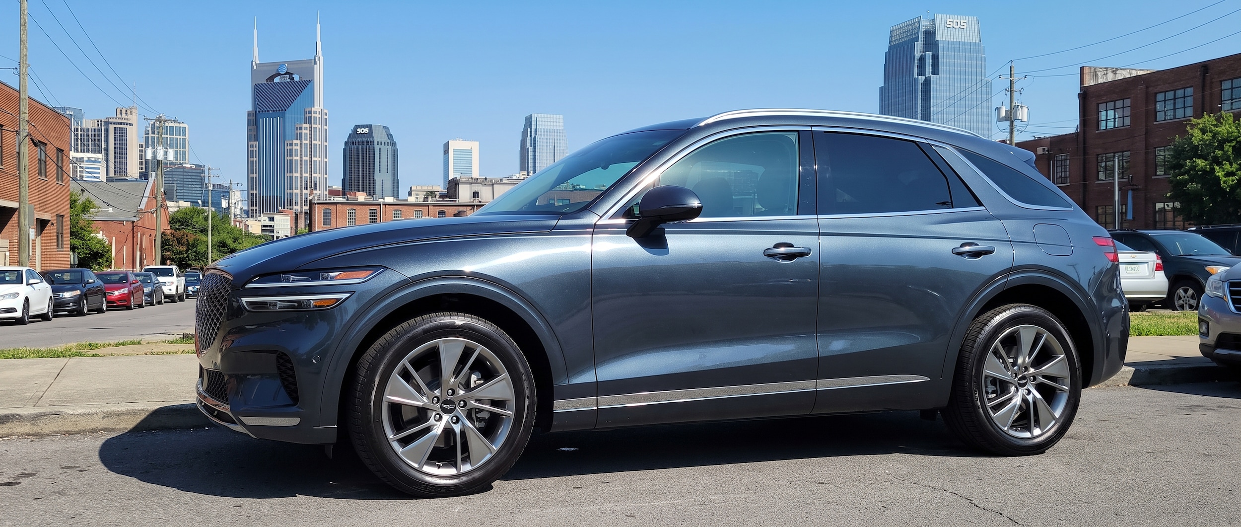 A 2026 Genesis Electrified GV70 in a deep metallic gray parked at an urban DC fast-charging station in Nashville, Tennessee on a clear summer afternoon, with the city skyline softly visible in the background