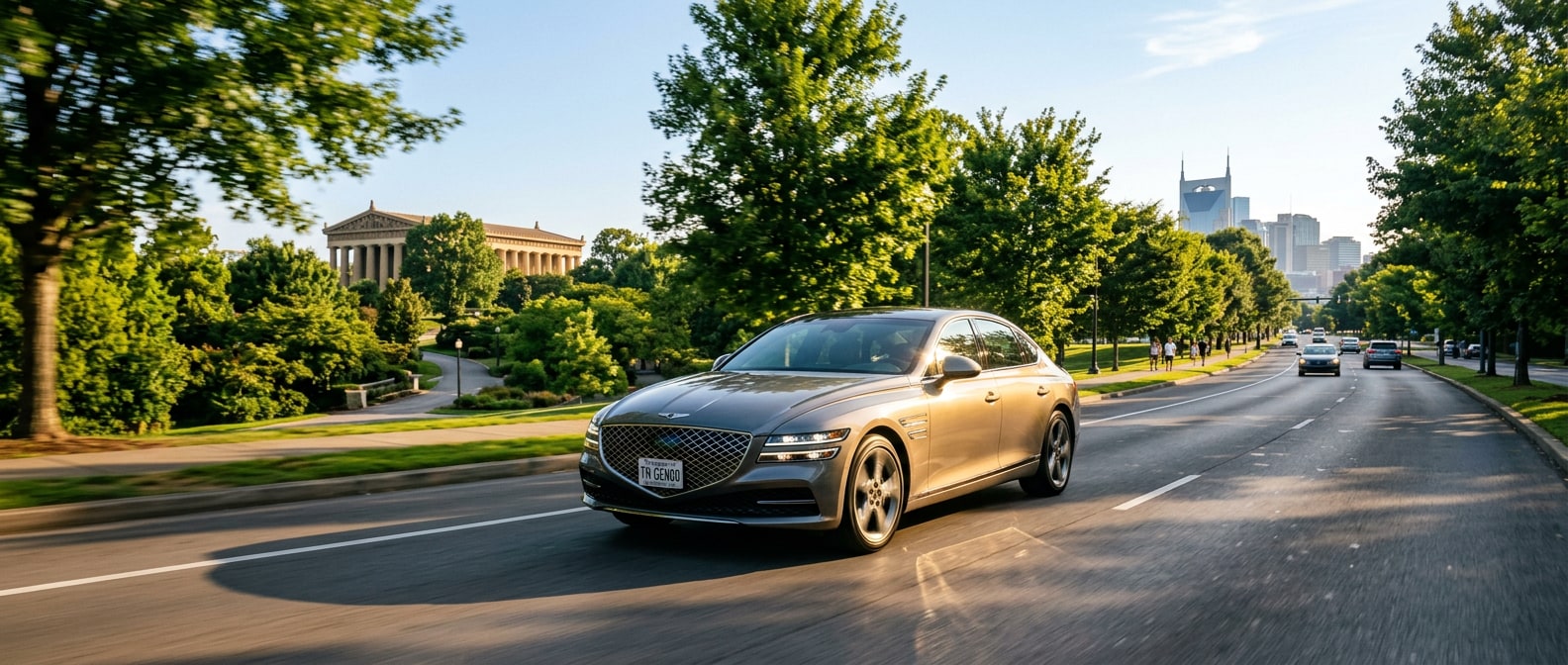 A 2026 Genesis G80 sedan in Makalu Gray moving through a tree-lined urban corridor on a bright late-summer Nashville morning, Centennial Park greenery visible in the background, warm golden light reflecting off the car's hood
