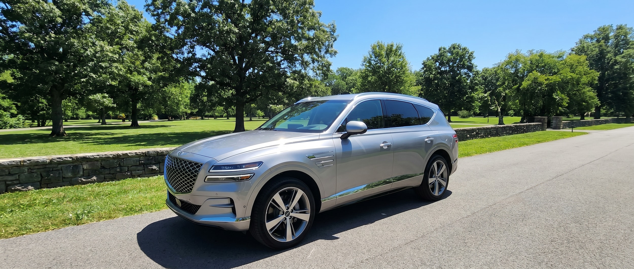 A silver Genesis GV80 parked near a lush green park in Nashville, Tennessee on a bright summer day, with sunlight reflecting off the hood and a clear blue sky overhead.