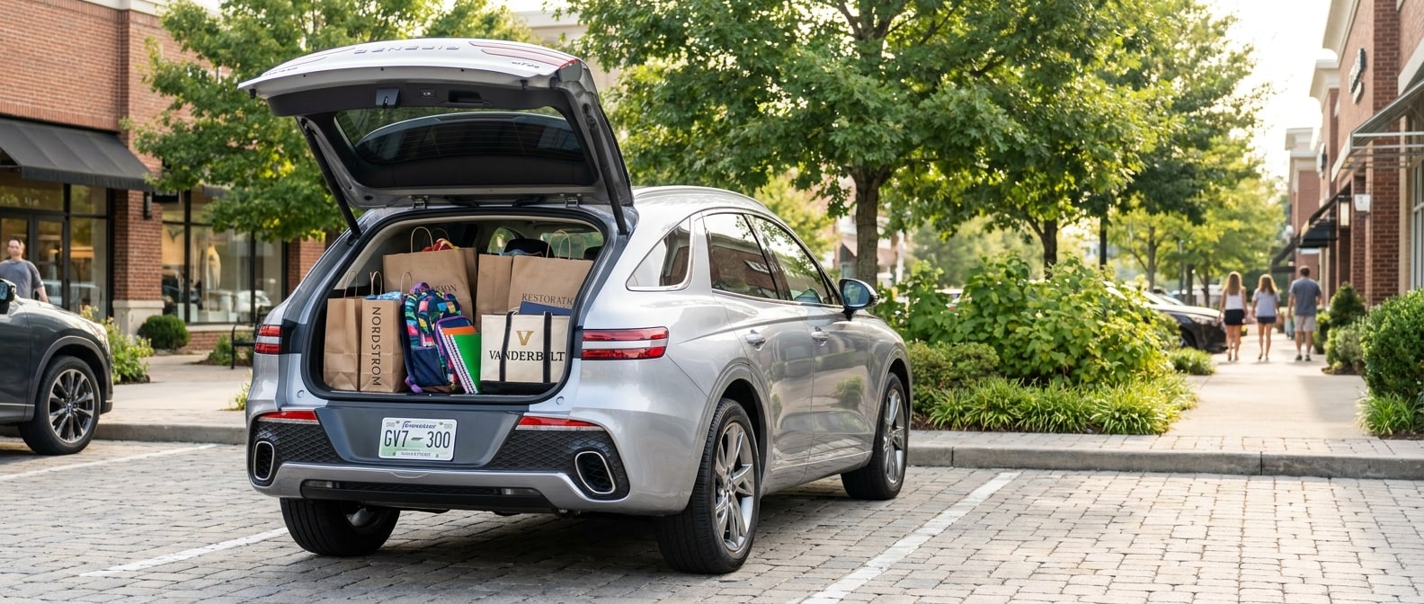 2026 Genesis GV70 in a polished silver or white finish parked in an upscale Nashville shopping center lot on a late-summer morning, back hatch open, school supplies and shopping bags loaded, green foliage visible in background suggesting the tree-lined streets of Green Hills