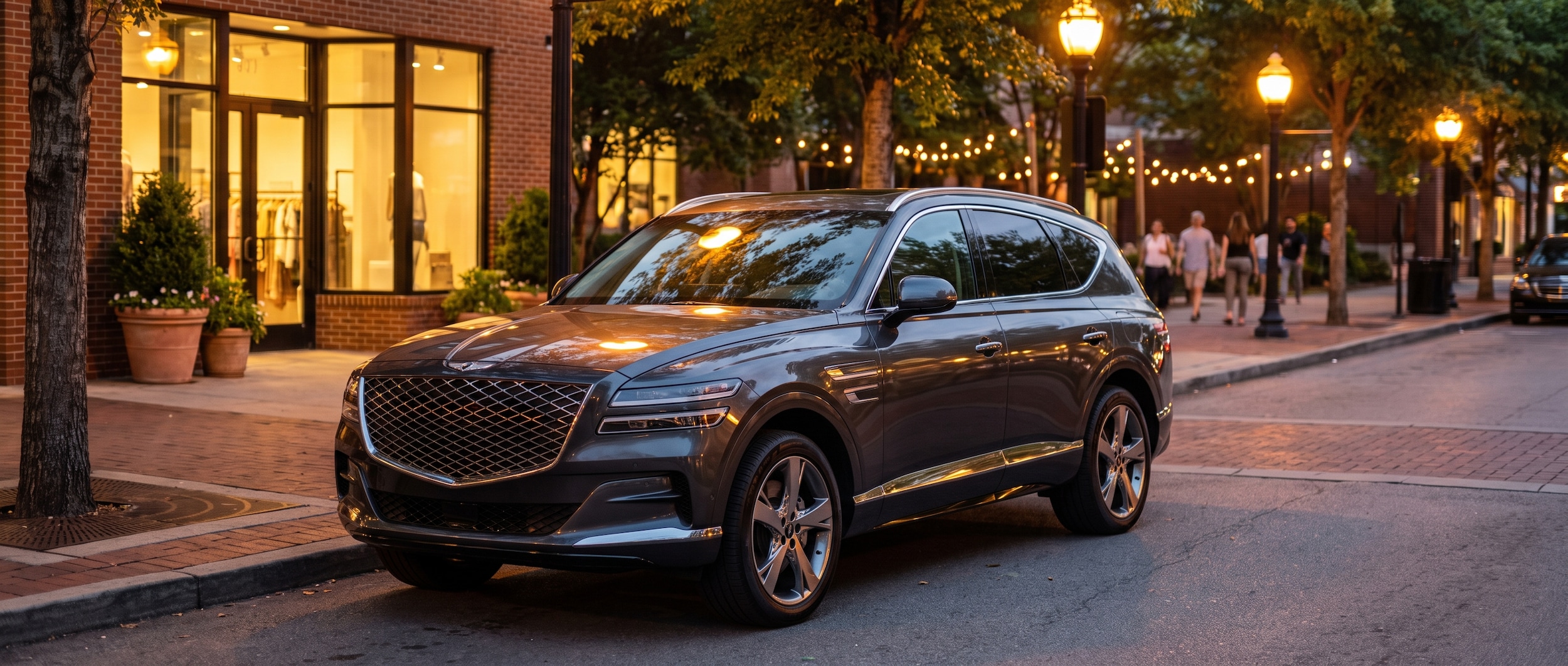 A 2027 Genesis GV80 in a dark, sophisticated exterior color parked along an upscale Nashville street at dusk, with warm streetlights reflecting off the hood and a softly lit boutique facade visible in the background, evoking a polished Green Hills evening out
