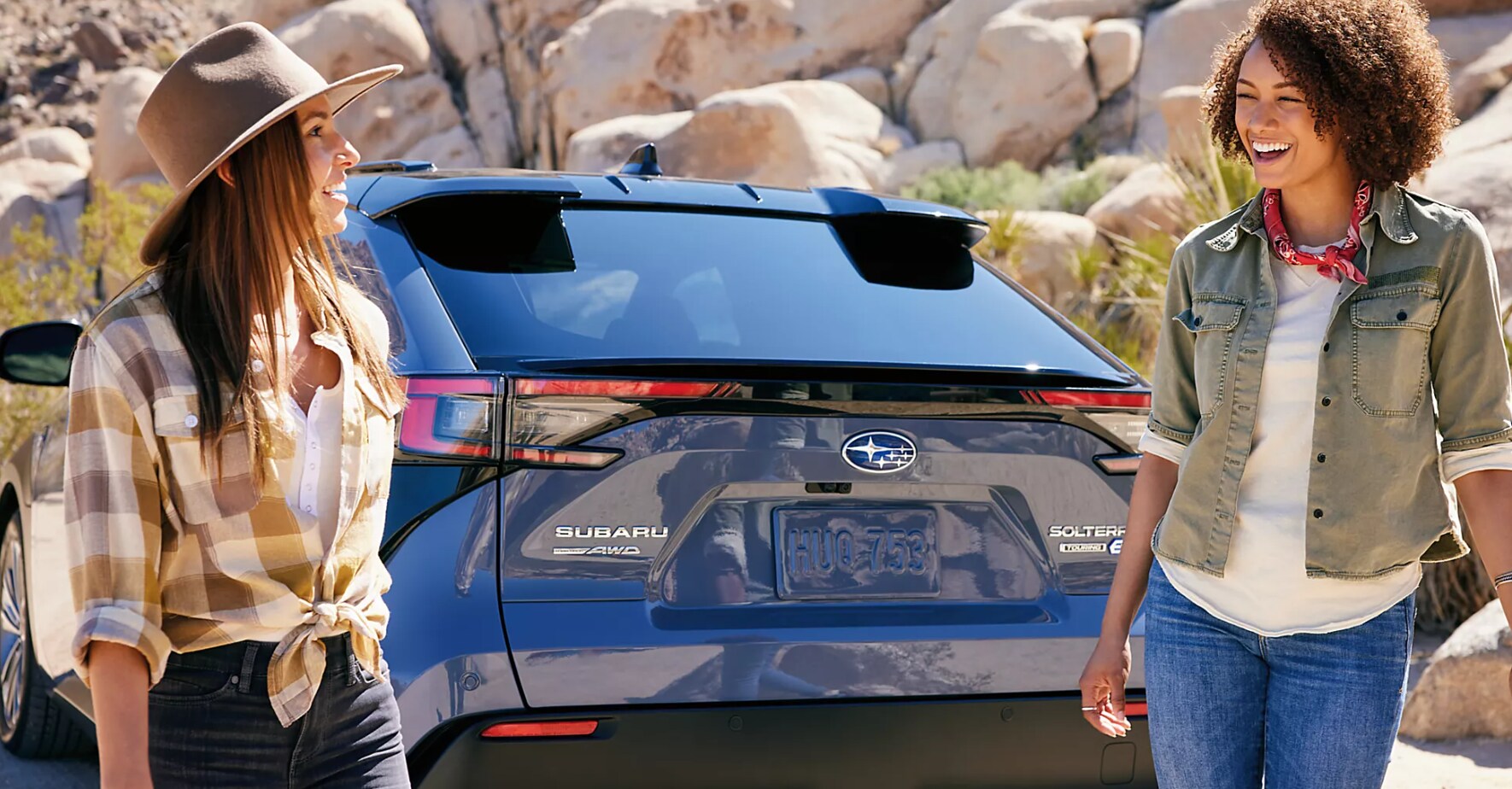 Two women chat in the foreground and the back of a blue 2023 Subaru Solterra is behind them