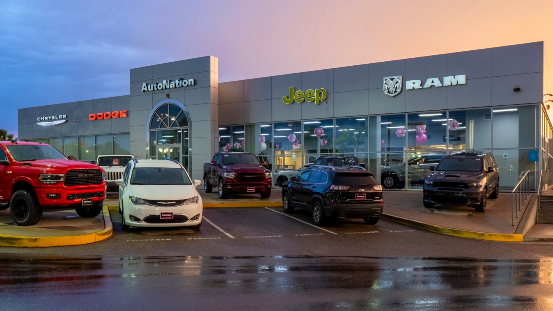 Exterior view of AutoNation Chrysler Dodge Jeep Ram Southwest during a sunrise or sunset. There is a slightly cloudy sky and many vehicles parked near the grey building. Trees and greenery are visible around the parking lot and surrounding area.