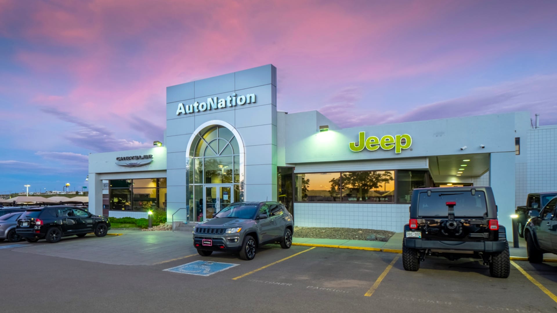 Exterior view of AutoNation Chrysler Jeep West during a sunrise or sunset. Many vehicles parked near the grey building. Trees and greenery are visible around the parking lot and surrounding area.