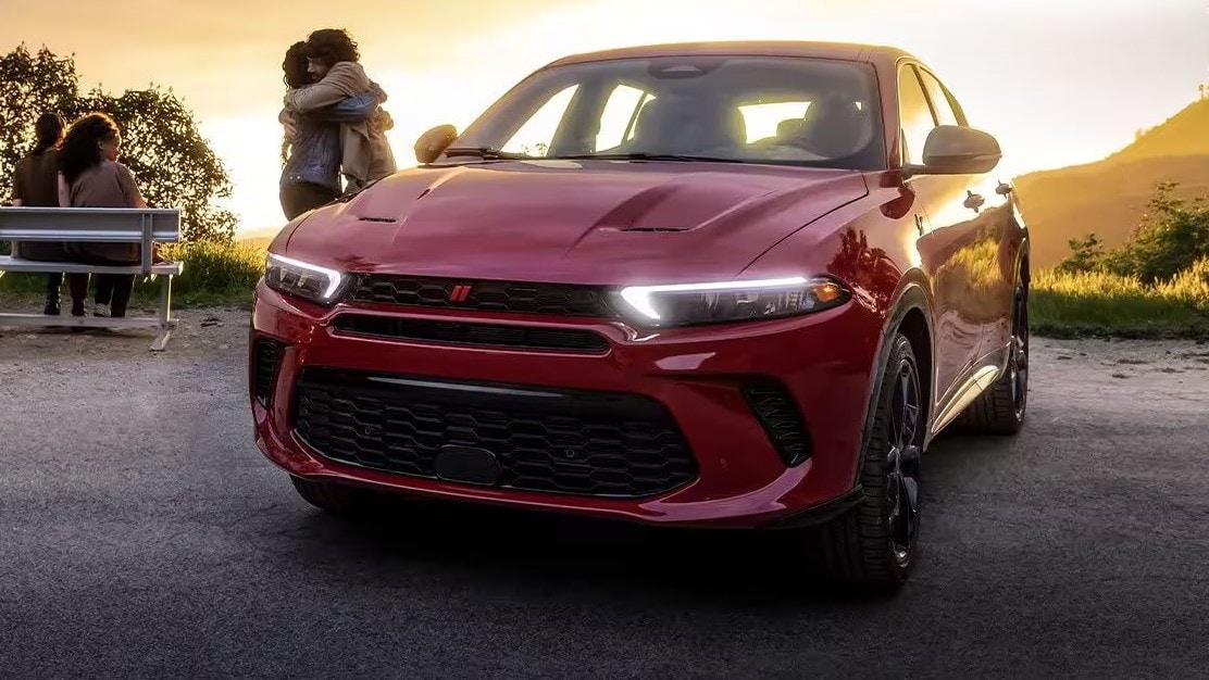 A red Dodge Hornet parked in a park in a sunset setting next to four people. Two of them are adults hugging next to the Dodge Hornet, while two others are seated on a bench off to the left.