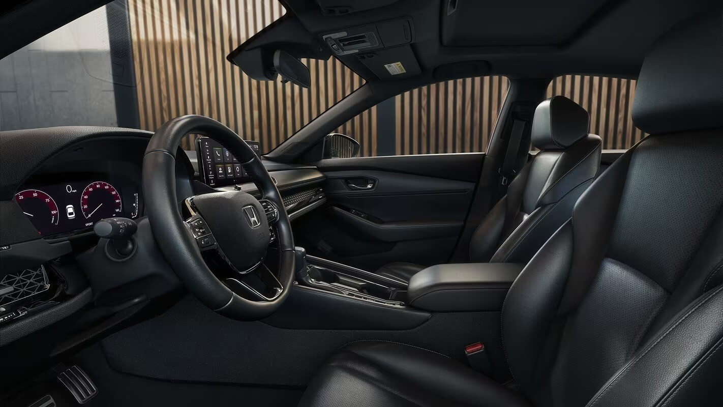  The front interior of the 2026 Accord from the driver's side, with a view of the seats, steering wheel, and gauges.