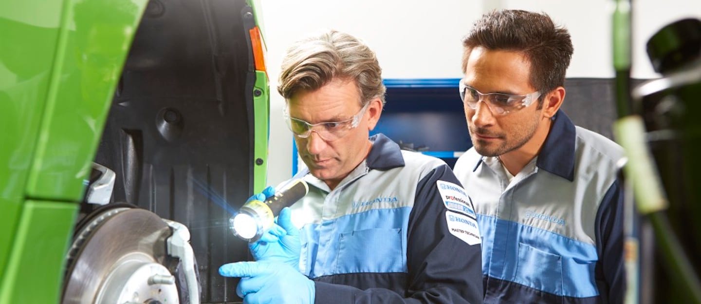 Two Honda service technicians wearing safety goggles inspect the breaks of a lime green vehicle.