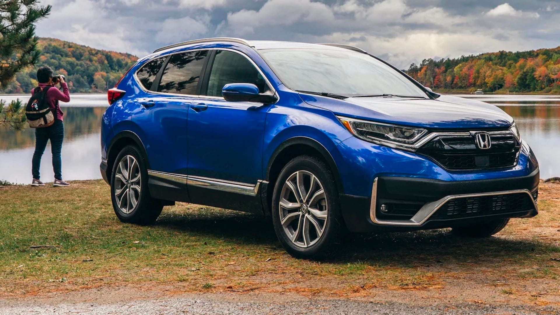 Blue Honda parked at the shore of a lake. A woman looks out towards the lake while taking a photo.
