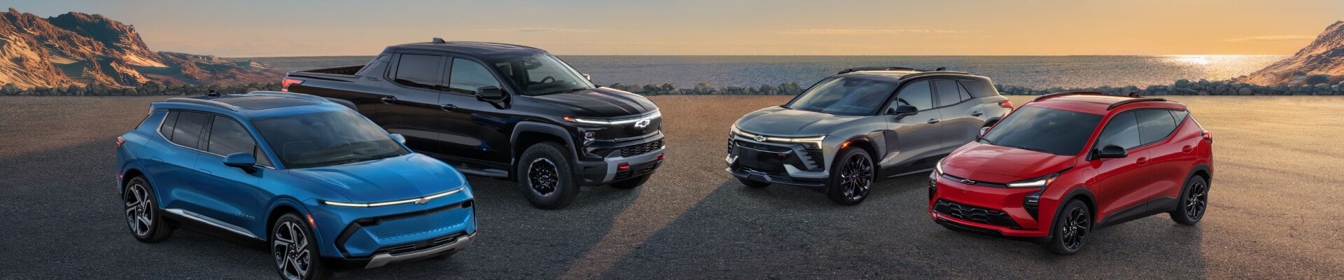 Front three-quarter view of blue, black, grey, and red Chevy EVs parked on asphalt near coastal mountains at sunset.