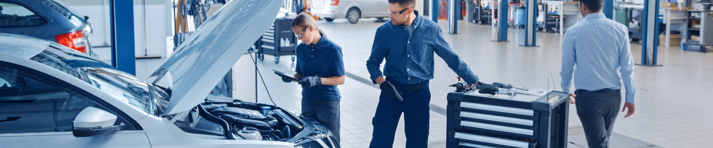 Mechanics in uniform work on a silver car with an open hood in a clean and well-lit garage.