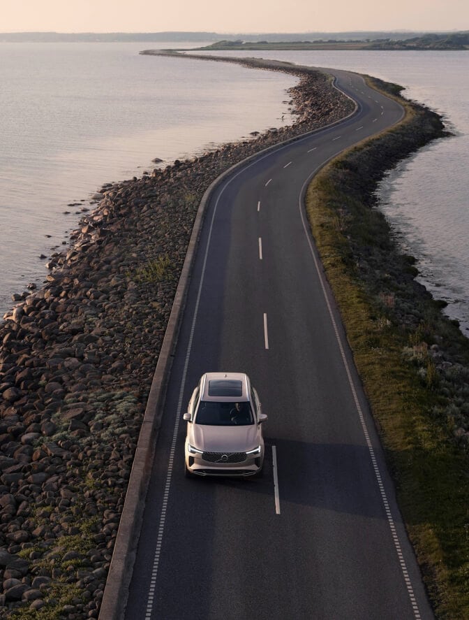 High-angle view of a beige Volvo SUV driving along a winding coastal road surrounded by water.