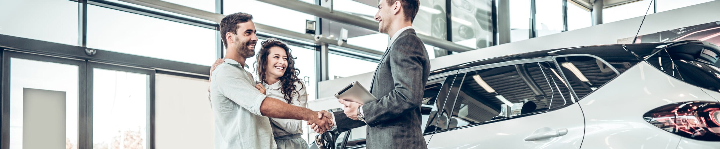 A smiling couple in a car showroom shakes hands with a salesperson beside a new SUV.