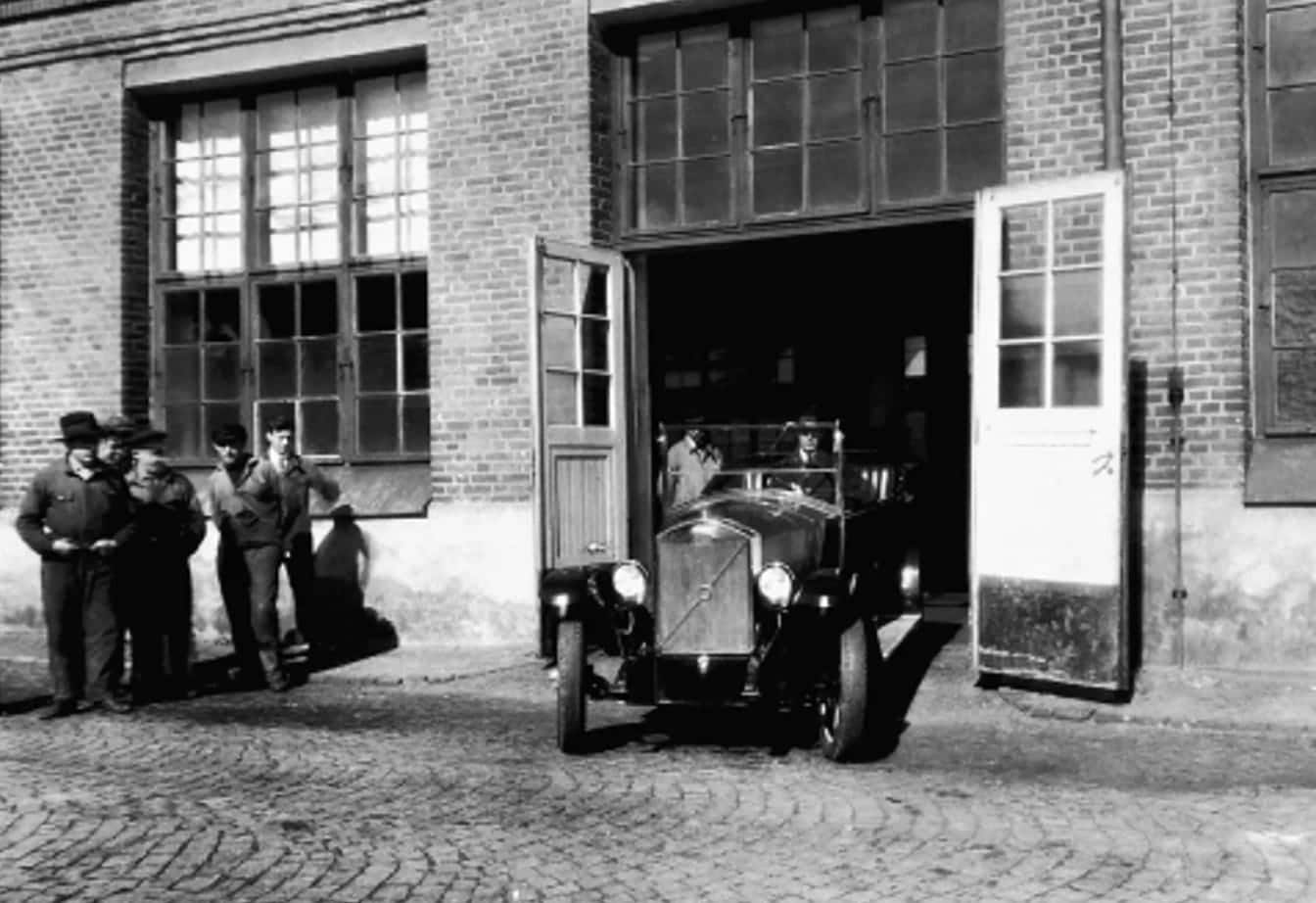 Historic black-and-white photo of an early Volvo vehicle leaving a factory with workers standing nearby.