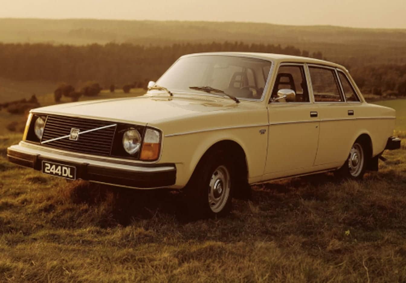 Vintage beige Volvo sedan parked in a grassy countryside landscape at sunset.