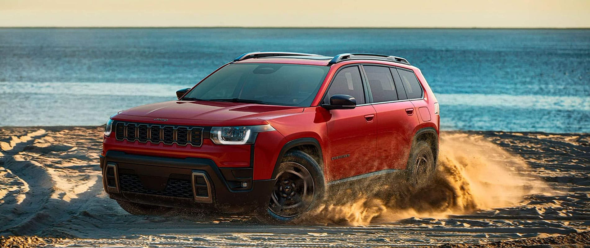A red Jeep Grand Cherokee kicking up sand as it drives on a beach.