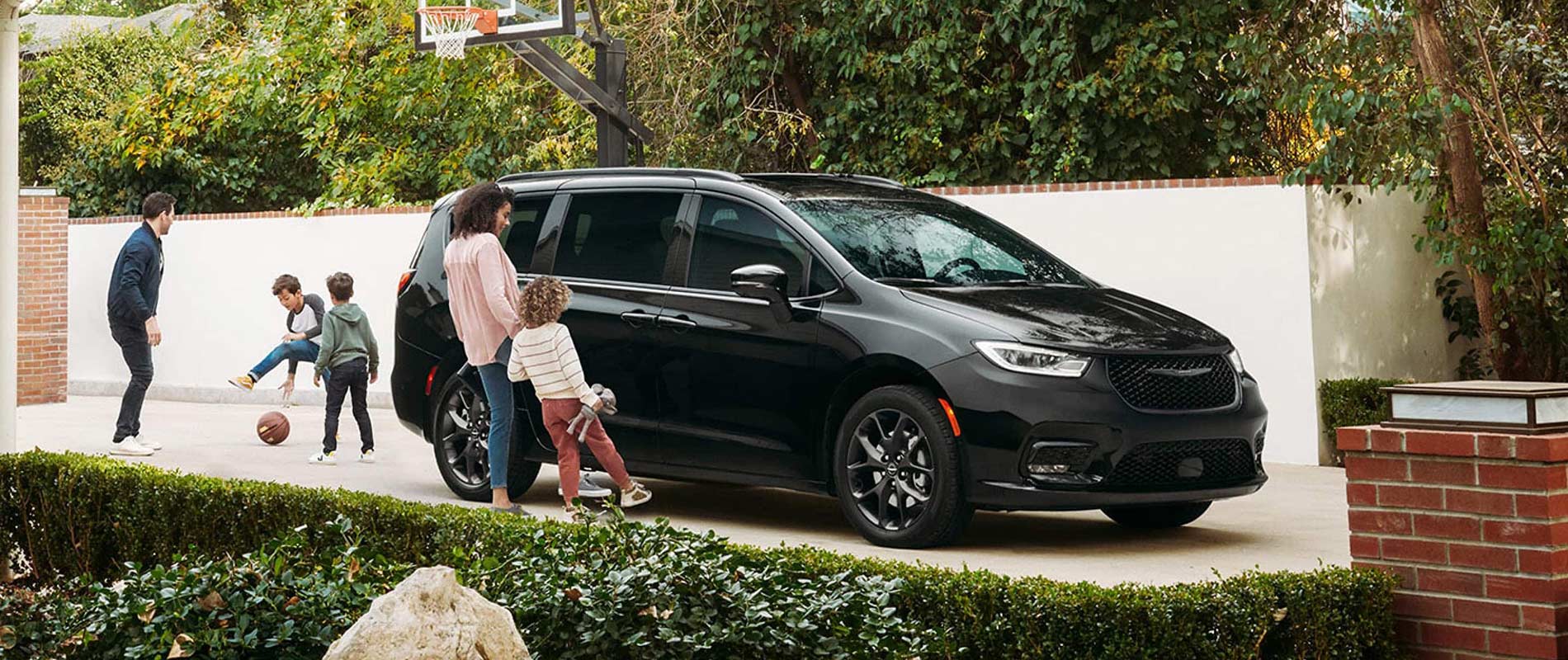 A black Chrysler Pacifica parked in a driveway by a basketball hoop, while a family plays around it.