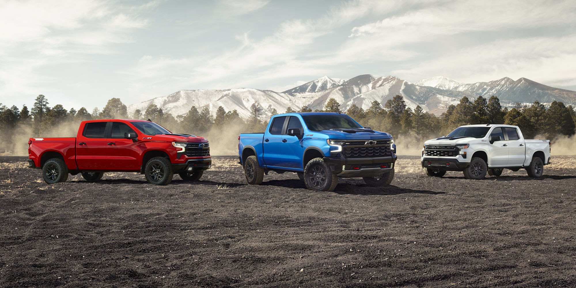 A trio of red, white, and blue Chevy Silverado models parked on dirt with a mountain backdrop