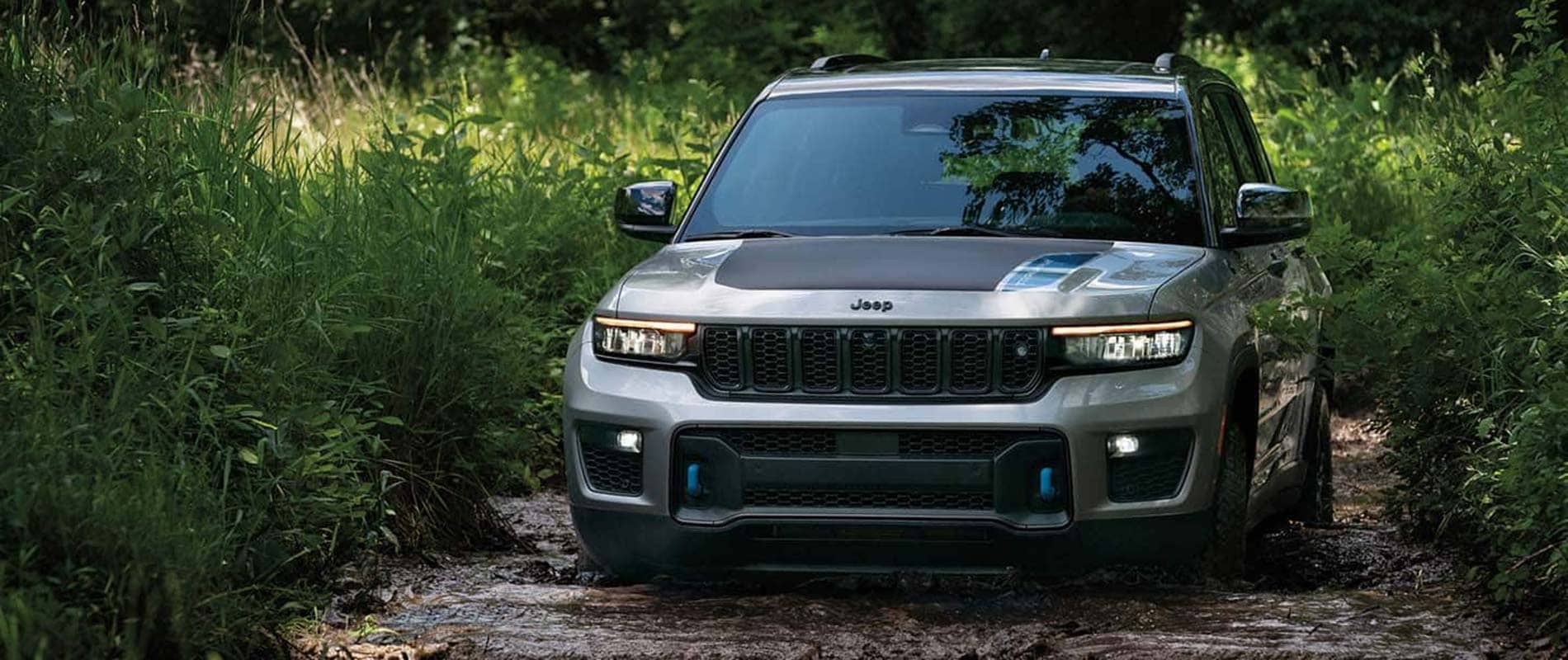 A silver Jeep Grand Cherokee driving over a muddy road in a forest.