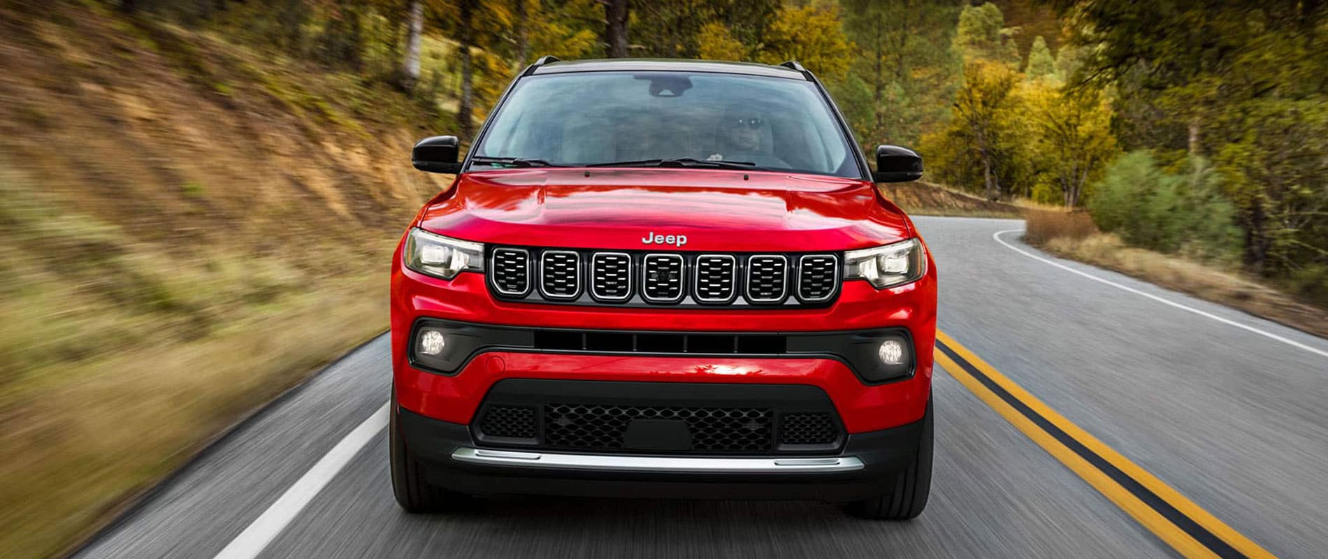A front view of a red Jeep Compass driving down the road past a forest.