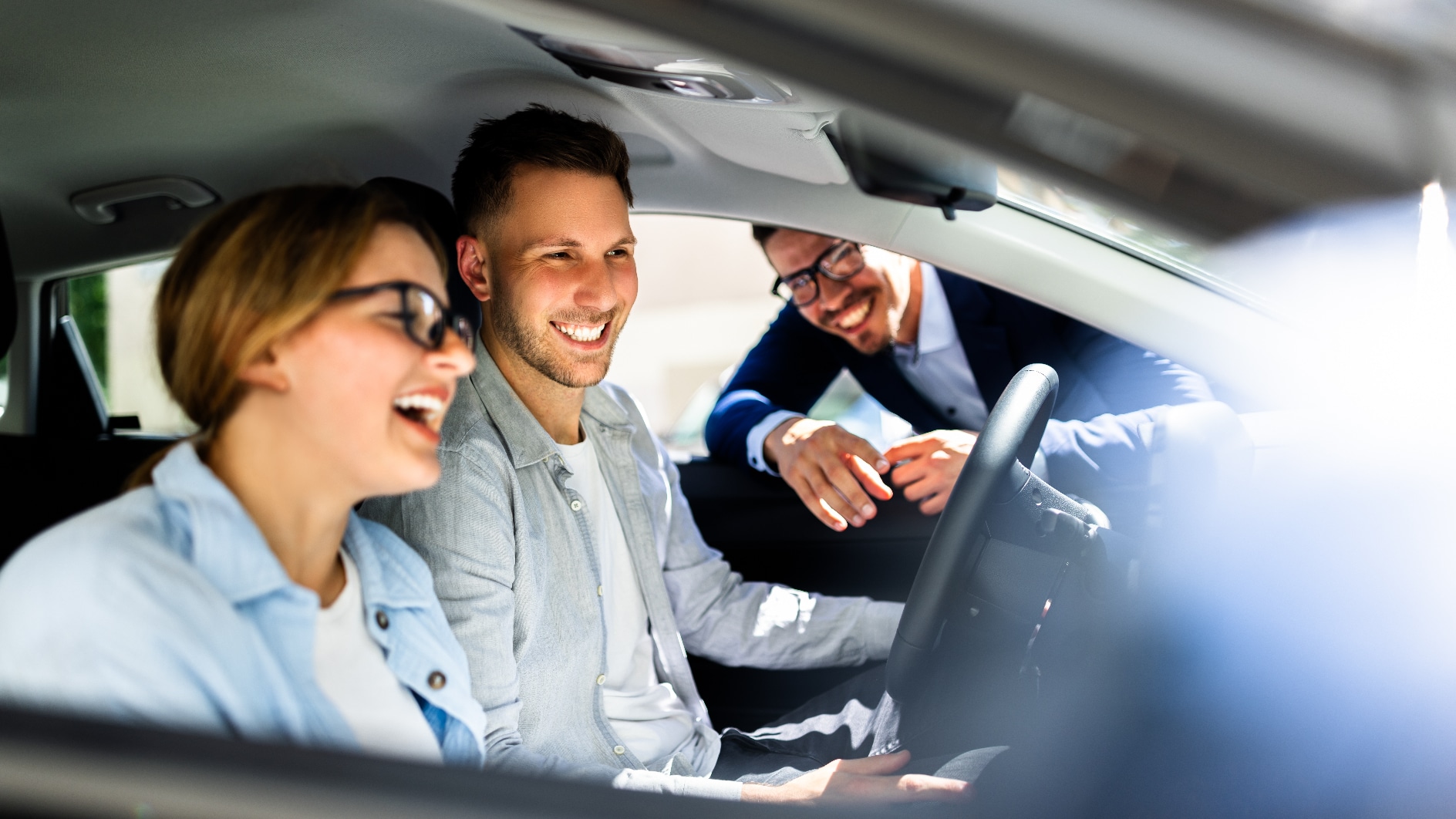 A couple behind the wheel of a car with a salesperson leaning through the window.