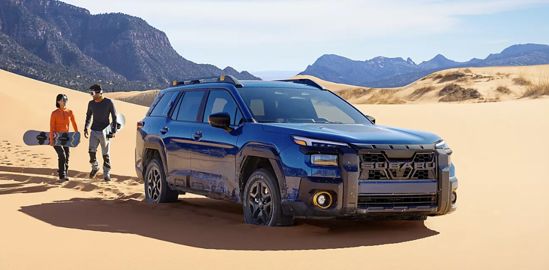 Blue 2026 Subaru Outback driving through desert sand with two person walking nearby