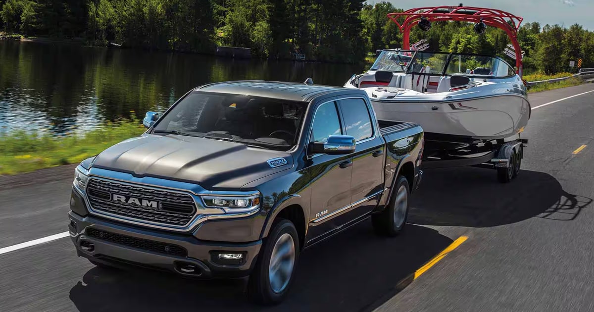 A 2024 Ram 1500 Limited Crew Cab with a silver exterior color towing a boat down a road by a lake.