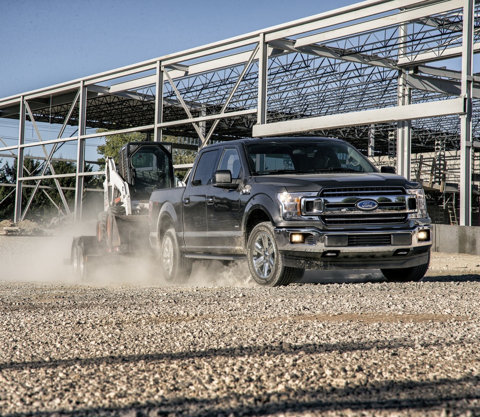 black Ford F-150 driving through a dusty construction area, towing a forklift on a trailer