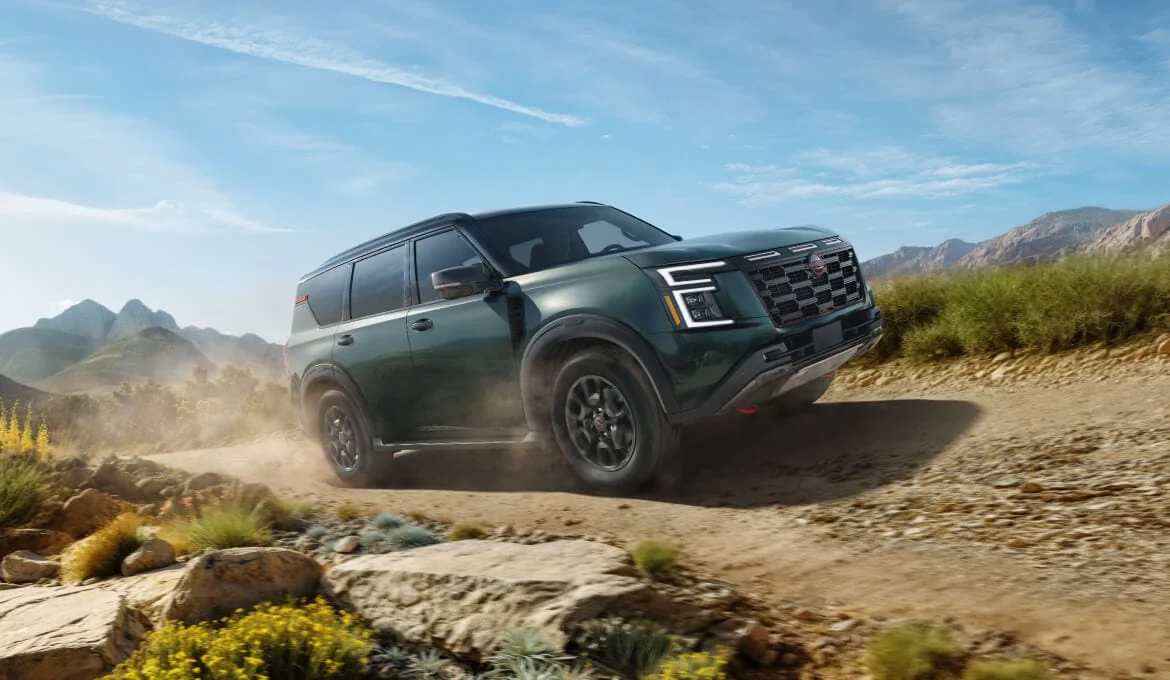 2027 Nissan Armada PRO-4X in dark green pulling up a dusty gravel track, with dust off the rear tire and mountains behind it