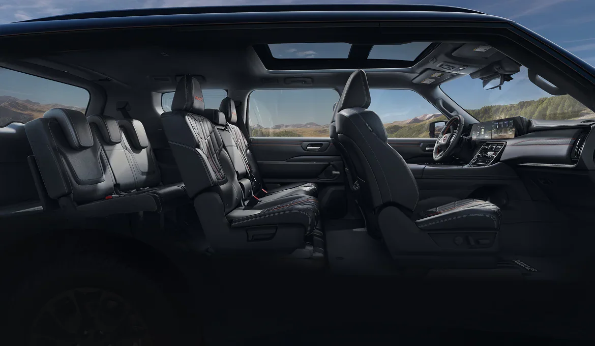 Side view through a 2027 Nissan Armada cabin showing all three rows of black seats, the panoramic roof overhead and the dual dashboard displays lit