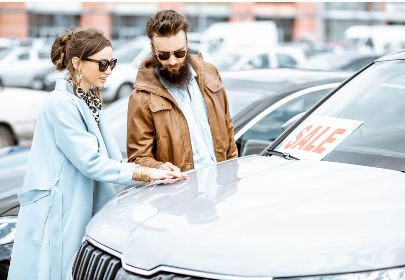 A couple wearing sunglasses inspects a used car with a sale sign in the window on a sunny day.