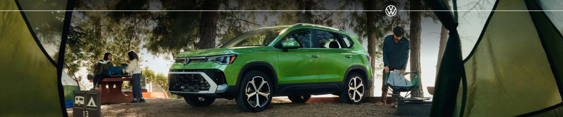 2027 Volkswagen Taos SEL in Bright Moss Green Metallic, displayed in front three-quarter view in a forest camping setting framed from inside a tent view.