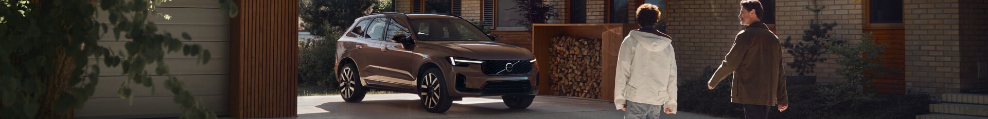 Front three-quarter view of a 2027 Volvo XC60 Ultra in Heather Bronze parked outside a house near two person walking.
