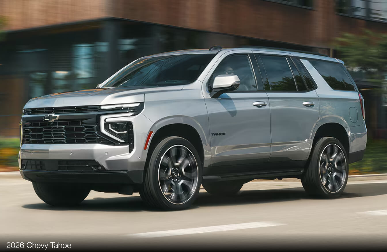 Exterior action shot of a silver 2026 Chevy Tahoe driving on a city road.