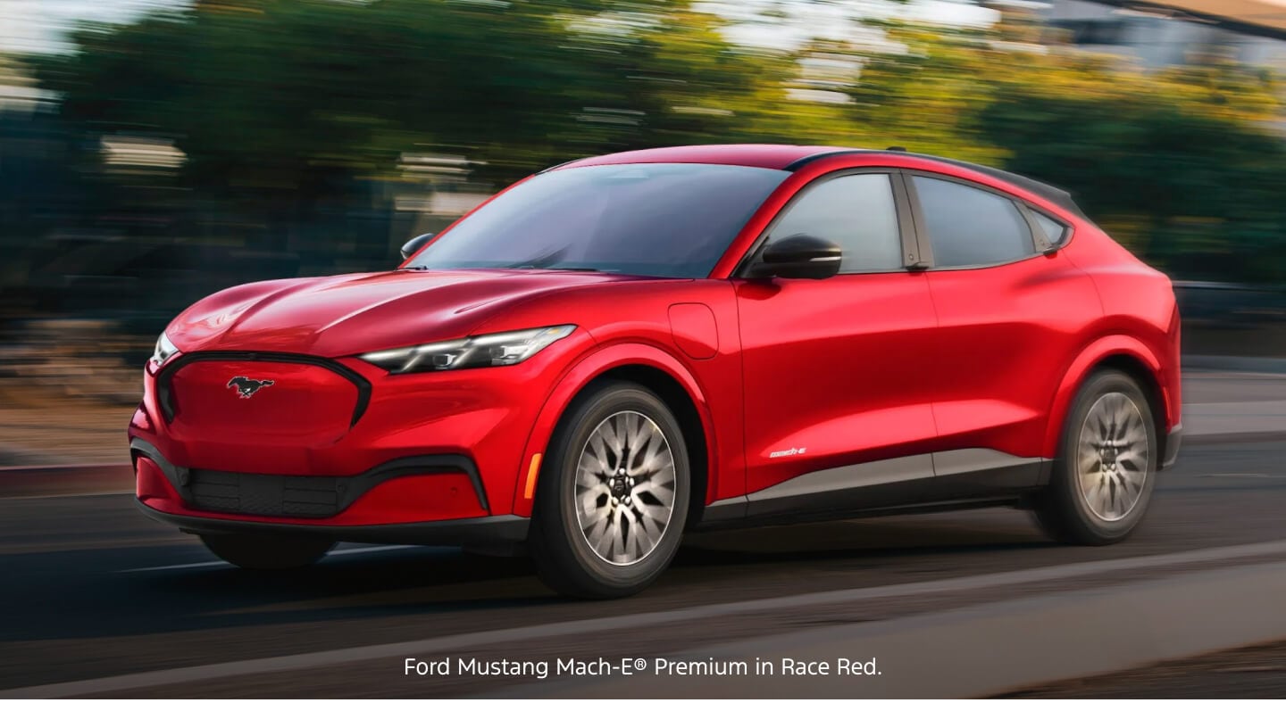 Three-quarter front view of a red Ford Mustang Mach-E Premium accelerating along a blurred urban street.