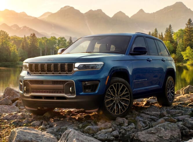2027 Jeep Grand Cherokee Overland in Fathom Blue, front three-quarter view on rocky terrain by a mountain lake at sunrise.