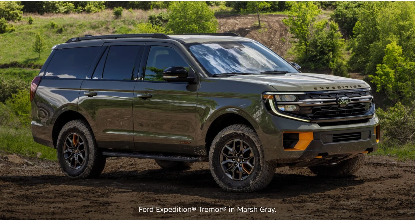 Three-quarter front view of a gray Ford Expedition Tremor parked on a muddy ground with green hills.