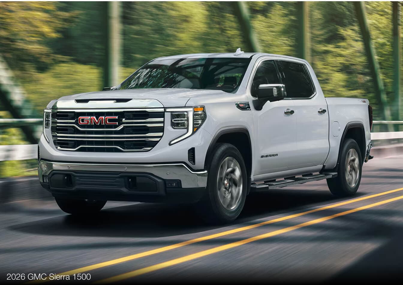 Exterior action shot of a silver 2026 GMC Sierra 1500 driving across a tree-lined bridge.