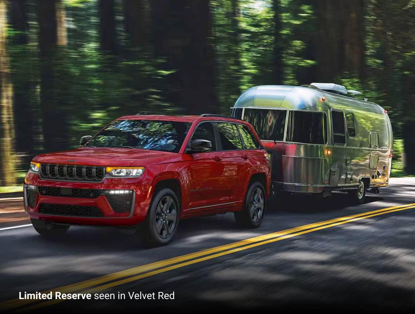2027 Jeep Grand Cherokee Limited Reserve in Velvet Red towing a silver travel trailer on a forest road.