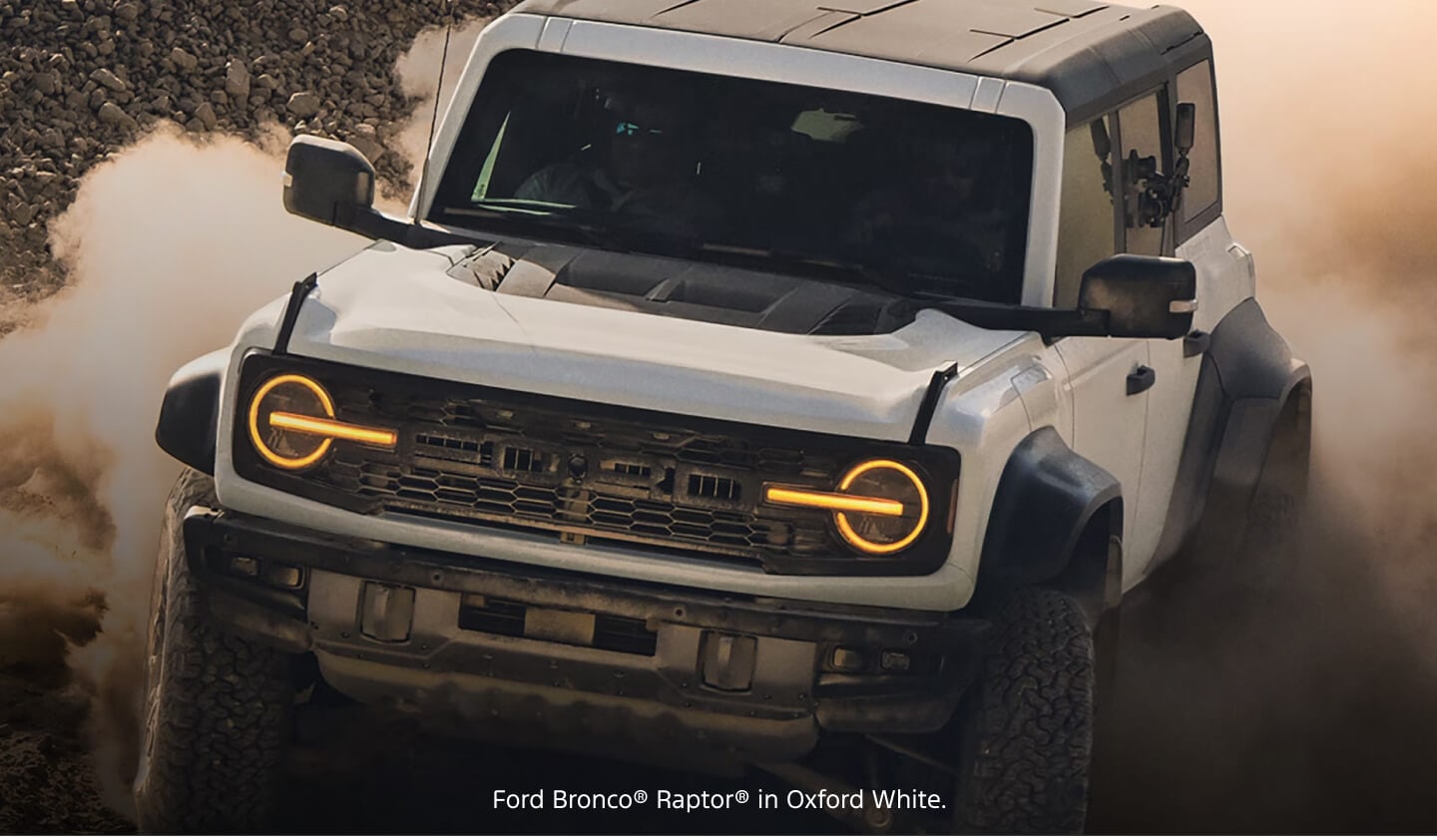 Front three-quarter view of a white Ford Bronco Raptor kicking up heavy dust in a rugged off-road environment.