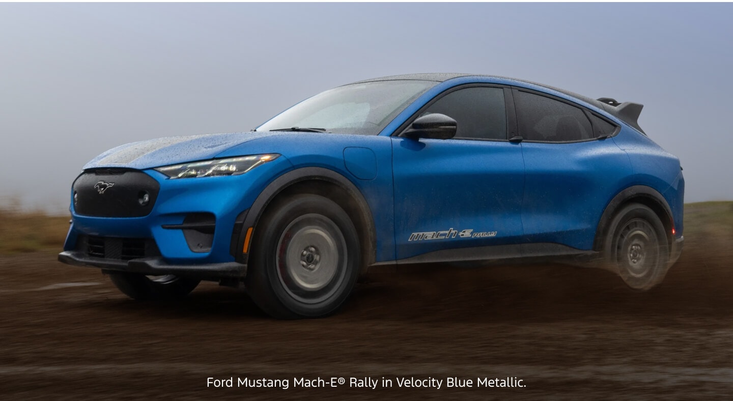 Low three-quarter view of a blue Ford Mustang Mach-E Rally drifting across a wet dirt course.