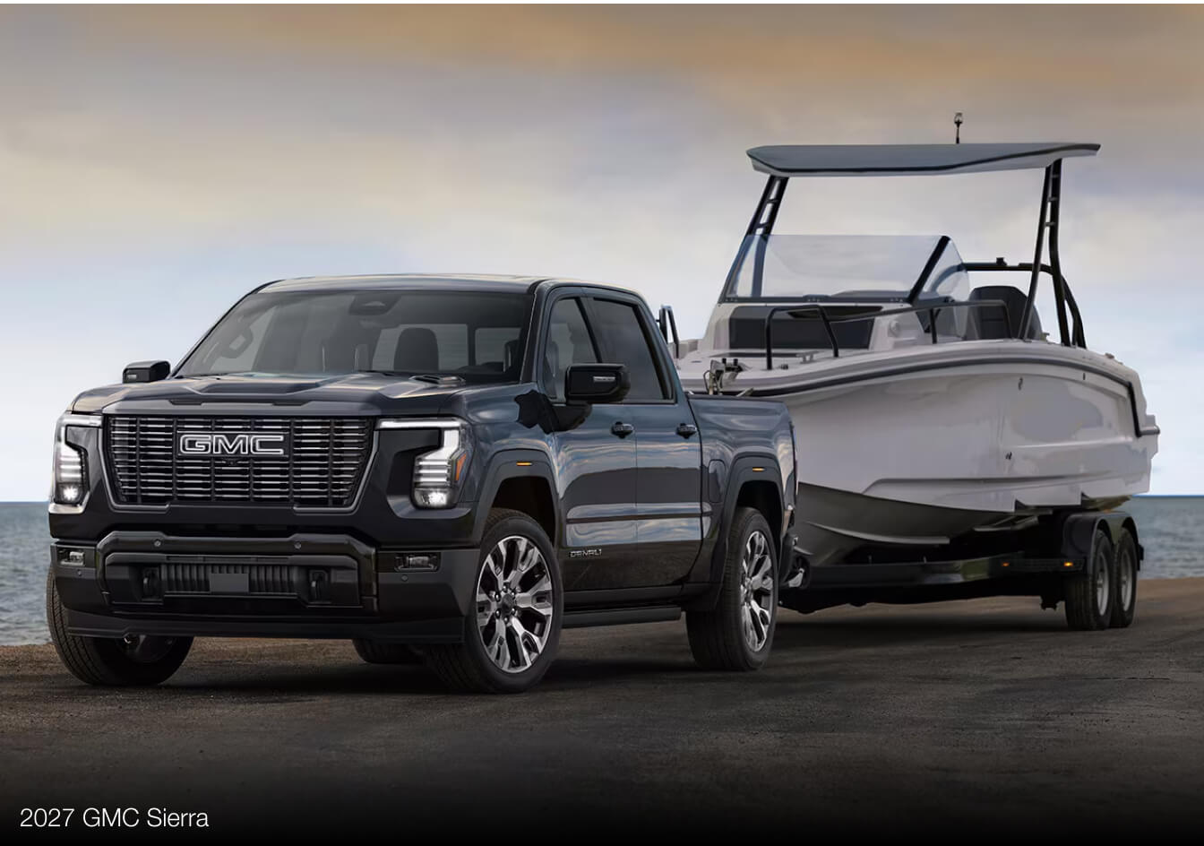 Exterior wide shot of a black 2027 GMC Sierra towing a boat near a body of water at dusk.