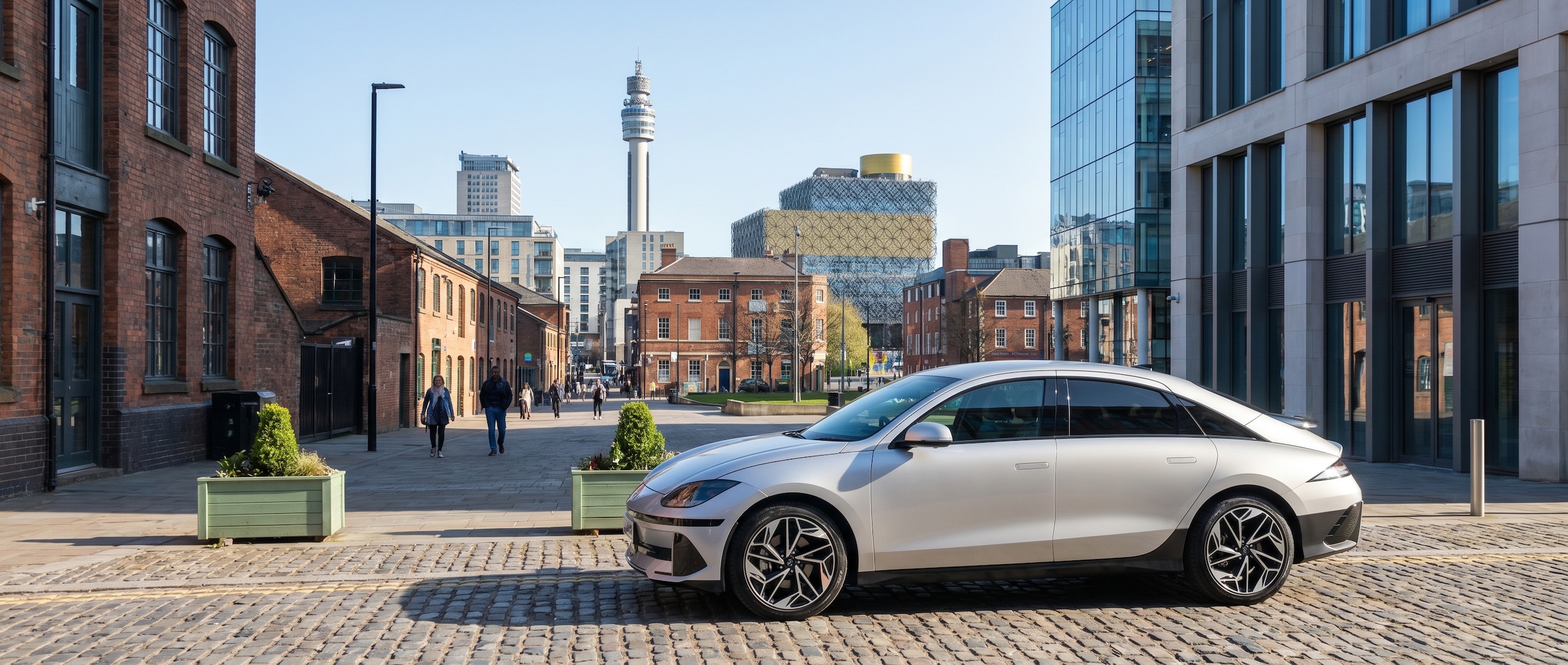 A sleek silver Hyundai IONIQ 6 parked in an urban Birmingham setting with the city skyline visible in the background on a clear afternoon, conveying a modern yet approachable feel.