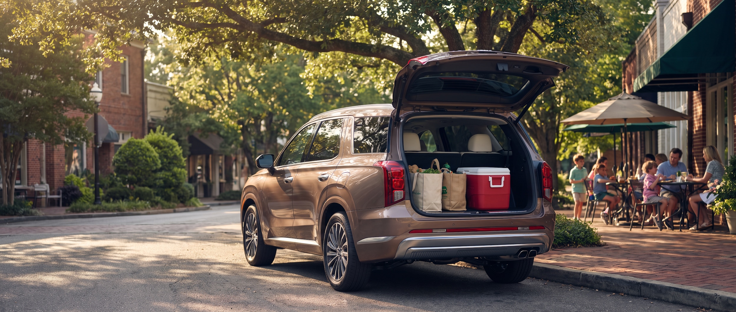 A 2026 Hyundai Palisade in a warm metallic color parked along a tree-lined Mountain Brook village street in summer, rear liftgate open with reusable bags and a picnic cooler visible, afternoon sunlight filtering through a dense tree canopy overhead, children and adults visible in the background near a sidewalk cafe.