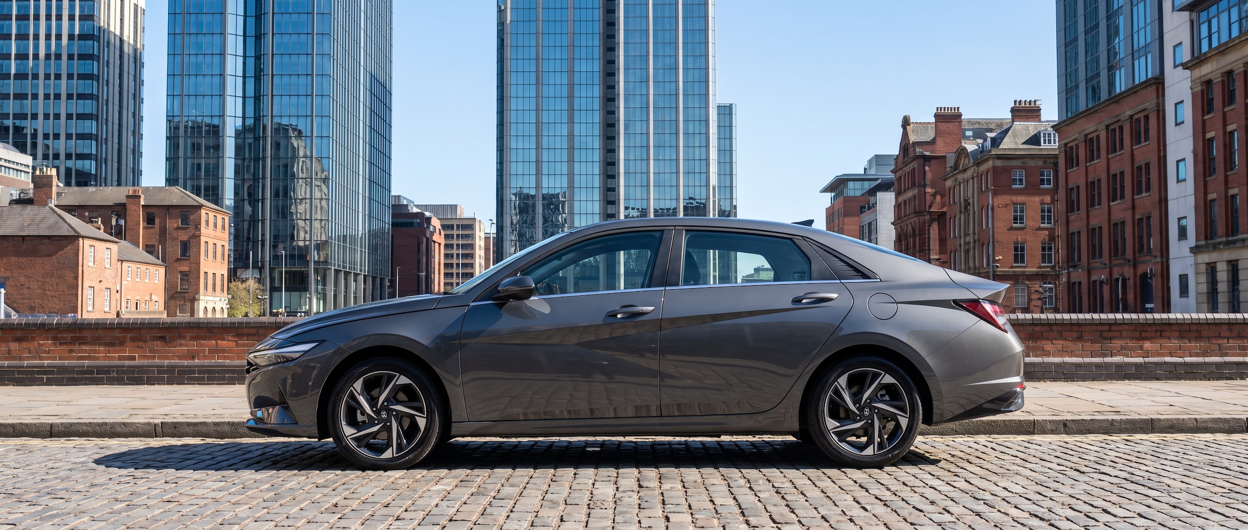 A 2026 Hyundai Elantra in a sleek modern color parked on a downtown Birmingham street, with the city's glass office towers visible in the background on a bright summer morning